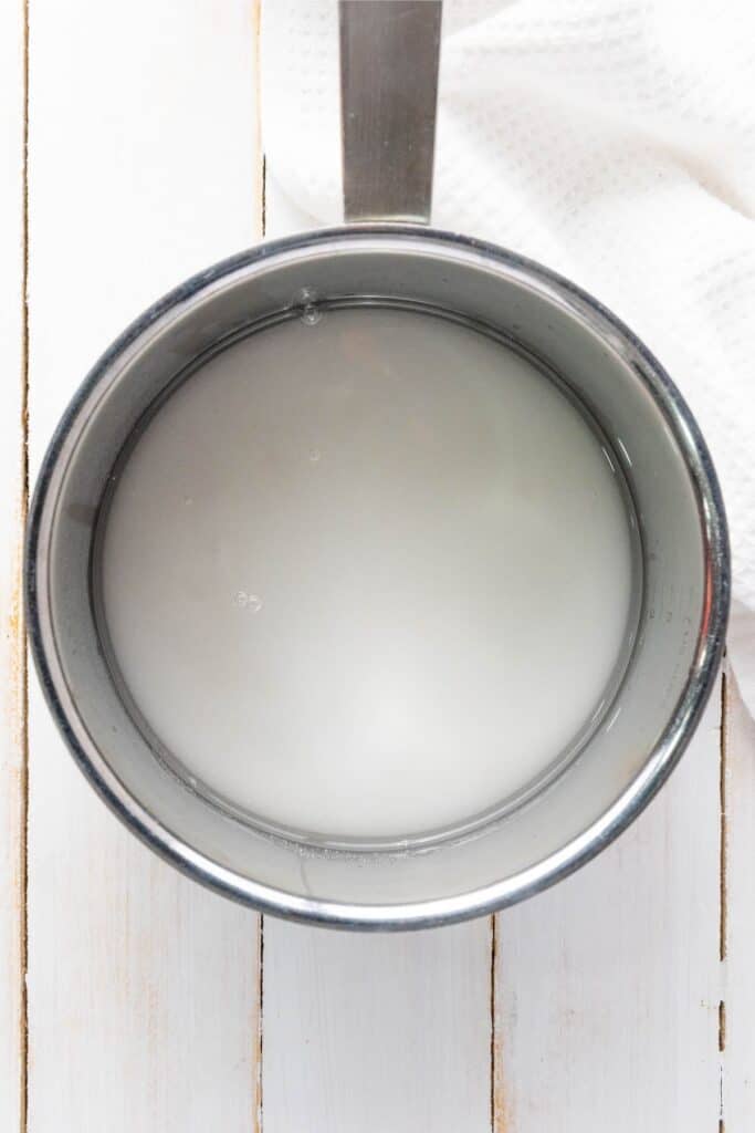 A stainless steel saucepan with cloudy liquid rests on a white wooden surface beside a textured cloth.