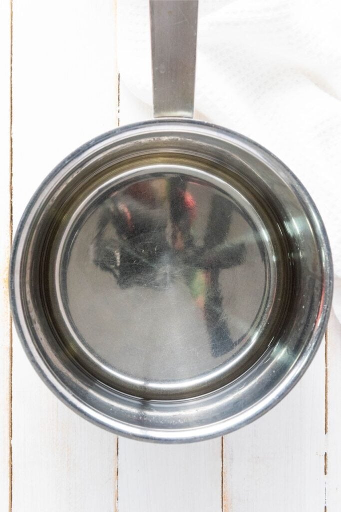 A stainless steel saucepan with clear water rests on a white wooden surface beside a folded white towel.